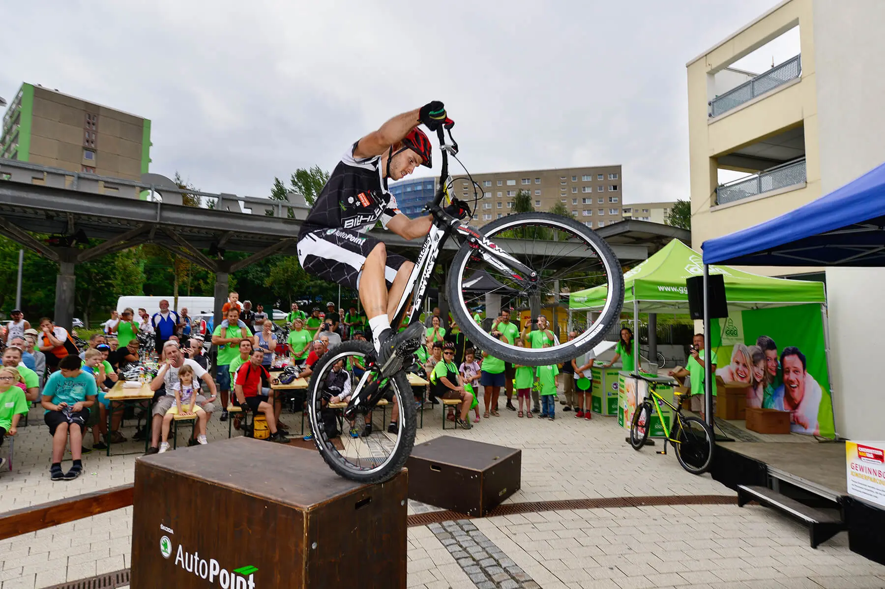 Ein Radfahrer vollführt während einer Trial Bike Show einen Stunt, indem er auf einem Mountainbike von einer Box zur anderen springt, während eine Menschenmenge in grünen Hemden das Ereignis im Freien in einer städtischen Umgebung beobachtet.