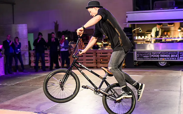 Ein Mann mit schwarzer Mütze und schwarzem Outfit führt in einer Halle einen Trick auf einem BMX-Rad vor, wobei er das Vorderrad wie ein Breakdancer vom Boden abhebt. Die Leute im Hintergrund schauen bei violettem Licht zu.