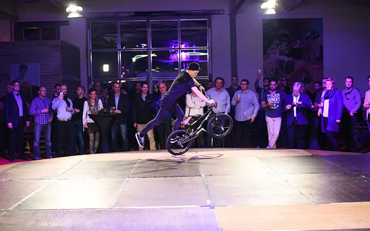 Ein BMX-Fahrer führt in der Mitte eines großen Raums einen Trick auf einem Rad vor und zieht die Menge wie ein Breakdancer in seinen Bann, während Jubel ausbricht und Scheinwerfer die energiegeladene Szene beleuchten.