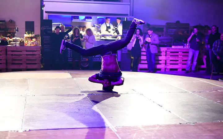 Ein Breakdancer balanciert kopfüber mit ausgestreckten Beinen in einer V-Form auf einer Tanzfläche, während die Leute im Hintergrund unter bunten Lichtern zusehen und jubeln.