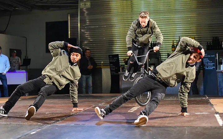 Drei Darsteller in passenden Jacken erhellen die Bühne; zwei sind Breakdancer, die dynamische Bodenbewegungen zeigen, während der dritte zwischen ihnen auf einem BMX-Rad fährt. Ein engagiertes Publikum schaut aus dem Hintergrund zu.