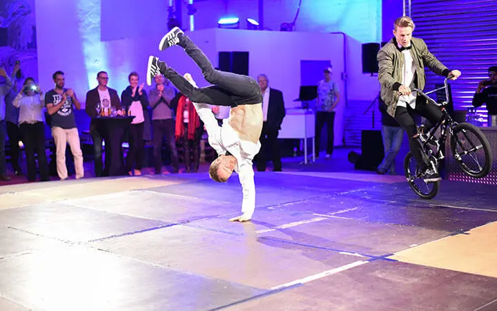 Ein Breakdancer in einem weißen Hemd führt einen einhändigen Handstand aus, während ein anderer Mann in der Nähe auf einem BMX-Rad fährt. Eine Gruppe formell gekleideter Menschen schaut zu und applaudiert in einer hell erleuchteten Halle.