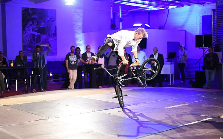 Ein Mann vollführt einen Trick auf einem BMX-Rad in einer Halle unter lila und blauem Licht. Er hebt das Hinterrad des Fahrrads an, während er auf dem Vorderrad balanciert und dabei eine Breakdance-ähnliche Beweglichkeit zeigt. Mehrere Personen schauen im Hintergrund zu.