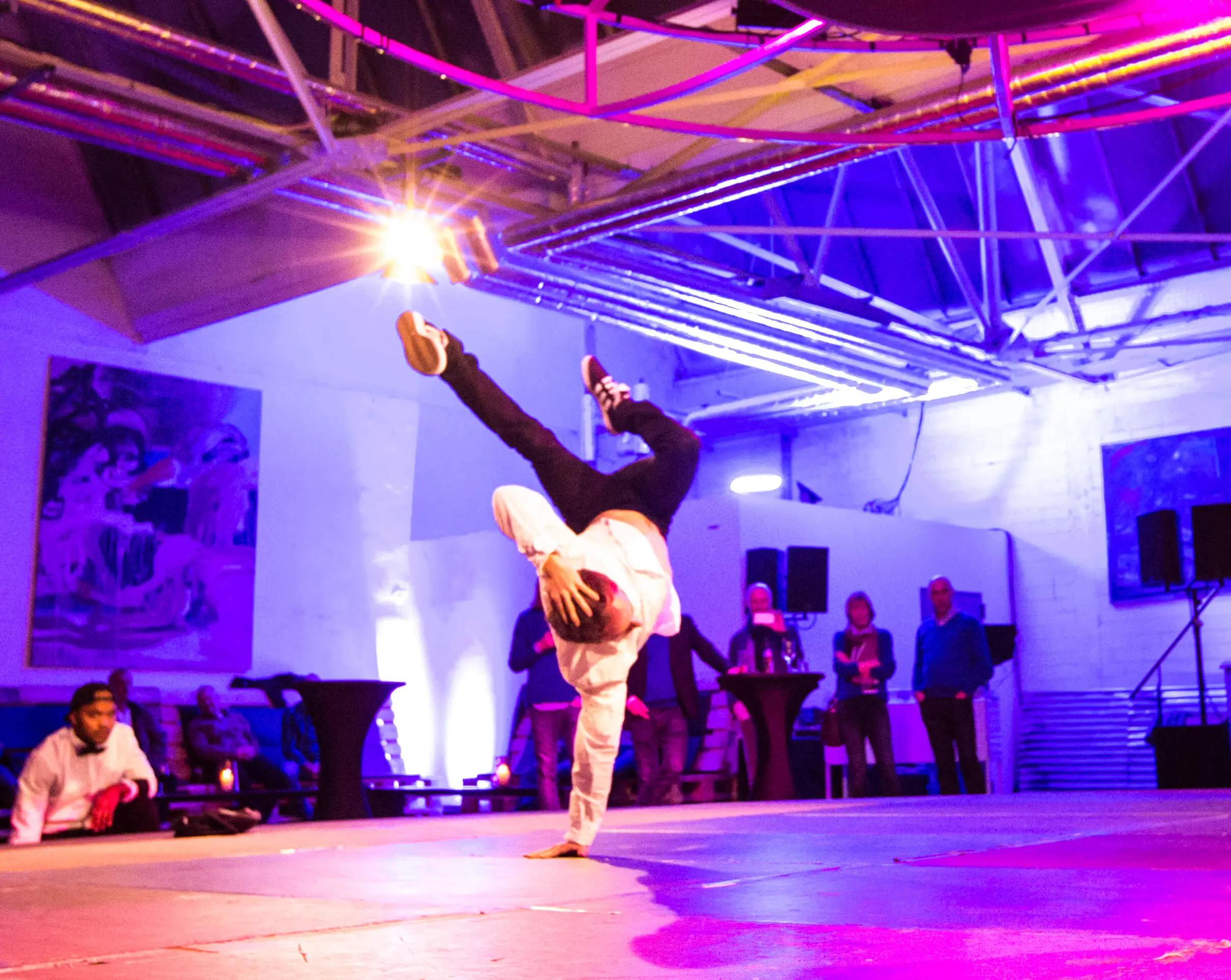 Ein Breakdancer balanciert in einem einhändigen Handstand auf der Bühne unter bunten Lichtern und fesselt das Publikum im Hintergrund eines Veranstaltungsortes im Industriestil.