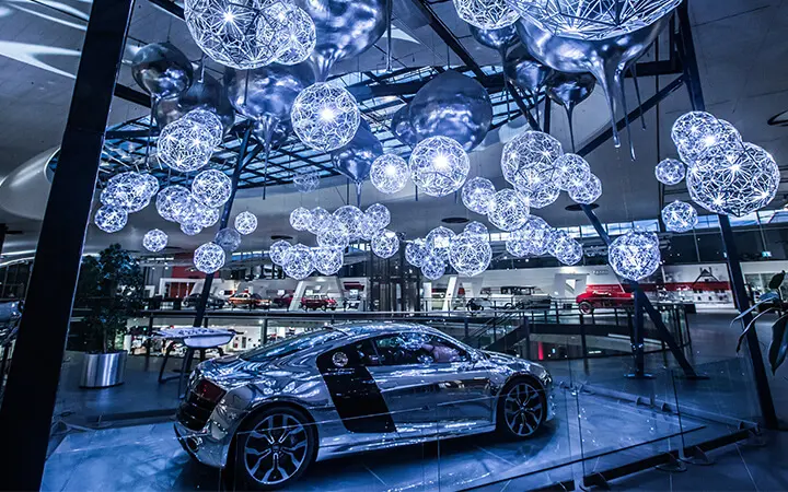 Ein silbern glänzender Sportwagen wird in einem Innenraum unter einer Deckeninstallation aus leuchtenden, kugelförmigen Lichtern und Metalldekorationen ausgestellt, die eine Urban-Dance-Atmosphäre heraufbeschwört, wobei im Hintergrund Spiegelungen und andere Autos zu sehen sind.