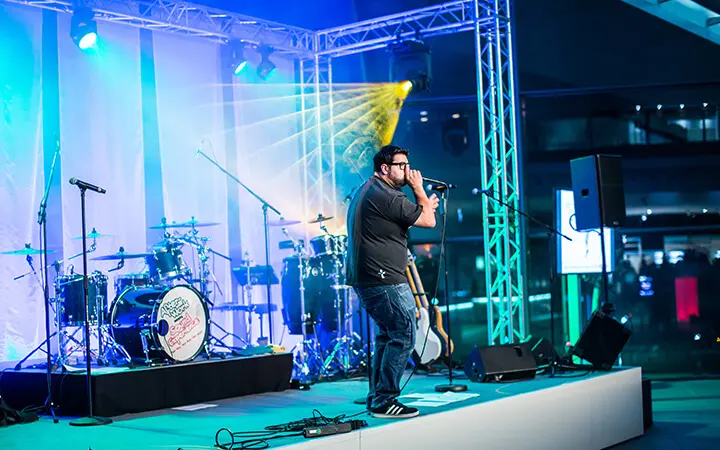 Ein Mann tritt auf einer hell erleuchteten Bühne mit einem Mikrofon auf. Hinter ihm stehen Musikinstrumente, darunter Schlagzeug und Gitarren. Bunte Lichter und ein moderner Veranstaltungsort in einer Halle verstärken die lebendige Atmosphäre dieser vom Urban Dance inspirierten Aufführung.