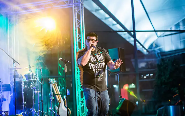 Ein Mann mit Brille und einem "Do The Positive Thing"-T-Shirt singt auf der Bühne in ein Mikrofon, umgeben von bunten Lichtern und Instrumenten, während Urban-Dance-Performer den beleuchteten Hintergrund beleben.