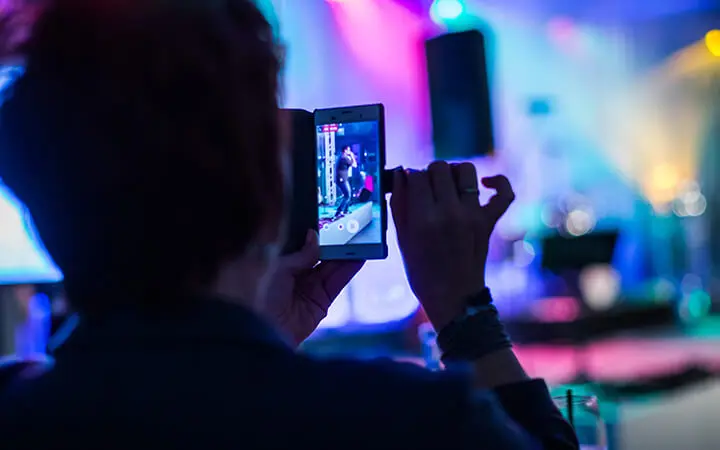 Eine Person hält ein Smartphone hoch, um eine Urban-Dance-Performance auf einer hell erleuchteten, farbenfrohen Bühne aufzunehmen oder zu fotografieren, wobei der Hintergrund unscharf ist.