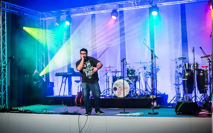 Ein Mann in einem schwarzen T-Shirt steht mit einem Mikrofon auf der Bühne und bringt Urban-Dance-Vibes in die Show. Hinter ihm leuchten bunte Bühnenlichter, und im Hintergrund sind Musikinstrumente wie Schlagzeug, Gitarren und Keyboards aufgebaut.