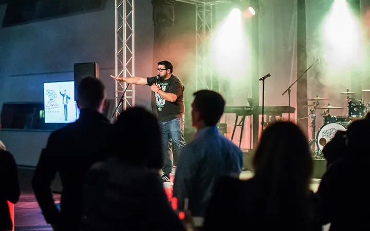 Ein Mann mit einem Mikrofon steht auf der Bühne einer Urban-Dance-Veranstaltung in einer Halle und spricht zum Publikum, während Musikinstrumente und Bühnenbeleuchtung die Szene untermalen und die Menschen im Vordergrund zusehen.