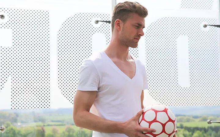 Ein Freestyler in einem weißen T-Shirt mit V-Ausschnitt hält einen weißen Fußball mit roten Mustern. Er steht im Freien, im Hintergrund sind eine gepunktete Glasscheibe und eine grüne Landschaft zu sehen.