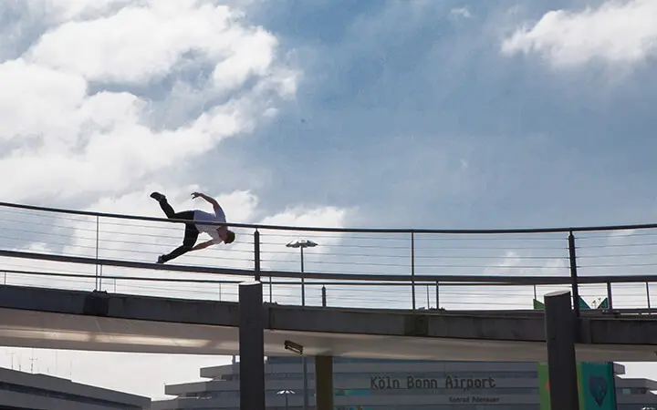 Ein Freestyler befindet sich in der Luft und springt über ein Metallgeländer auf einer Fußgängerbrücke in der Nähe des Flughafens Köln/Bonn, mit einem bewölkten Himmel im Hintergrund.