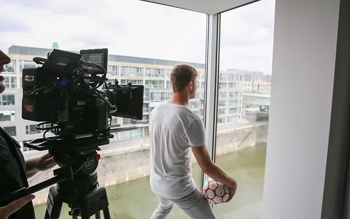 Ein Freestyler in weißer Kleidung hält einen Fußball und schaut aus einem großen Fenster, während ein Kameramann sie filmt. Draußen sind Gebäude und Wasser zu sehen.