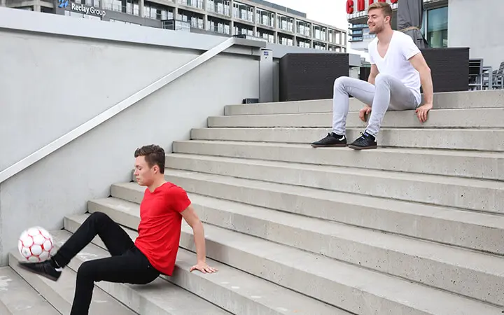 Zwei junge Männer stehen auf einer Betontreppe im Freien; einer im roten Hemd sitzt und zeigt seine Fußballkünste mit einem Ball, während der andere im weißen Hemd weiter oben sitzt und zusieht.