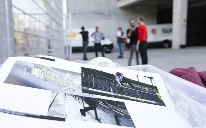 Eine Nahaufnahme von gedruckten Schwarz-Weiß-Skateboarding-Fotos, die den dynamischen Geist eines Freestylers einfangen, mit einer verschwommenen Gruppe von fünf Personen, die im Freien in der Nähe eines weißen Lieferwagens und eines Betongebäudes im Hintergrund stehen und sich unterhalten.