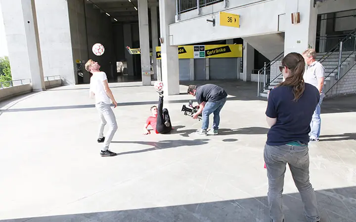 Vier Personen stehen, während ein Freestyler auf dem Rücken liegt und geschickt einen Fußball mit den Füßen balanciert. Ein Kameramann filmt das beeindruckende Kunststück in einer von Betonpfeilern und gelben Schildern gesäumten Stadionhalle.