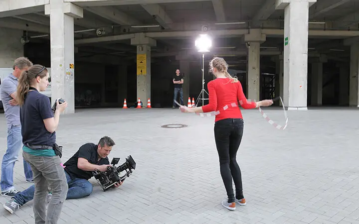 Eine Frau in einem roten Hemd springt Seil wie eine echte Freestylerin, während ein Filmteam sie aufnimmt. Drei Personen stehen in der Nähe, eine bedient eine Kamera in Bodennähe. Helle Lichter und Verkehrskegel unterstreichen die städtische Betonkulisse.
