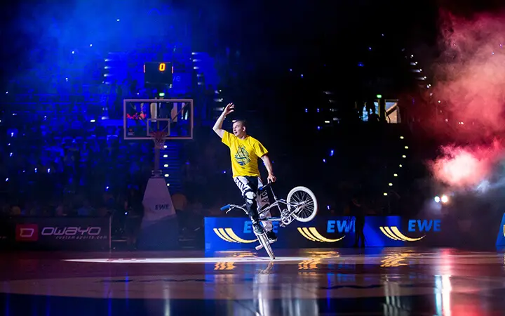 Ein BMX-Fahrer sorgt für Unterhaltung, indem er unter dramatischer Beleuchtung in einer Reithalle einen Stunt auf einem Rad vorführt. Er trägt ein gelbes Hemd und eine gemusterte Hose, im Hintergrund sind bunter Rauch und ein Publikum zu sehen.