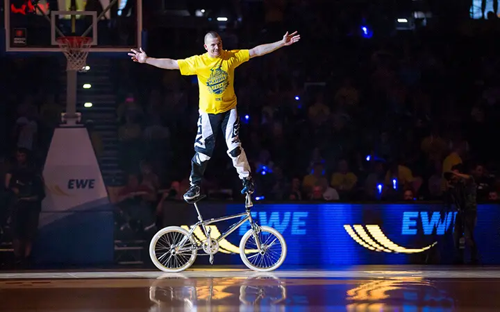 Ein Darsteller in einem gelben Hemd und einer schwarzen Hose balanciert mit ausgestreckten Armen auf dem Sattel und Lenker eines BMX-Rads während einer Show in einer Indoor-Entertainment-Arena.