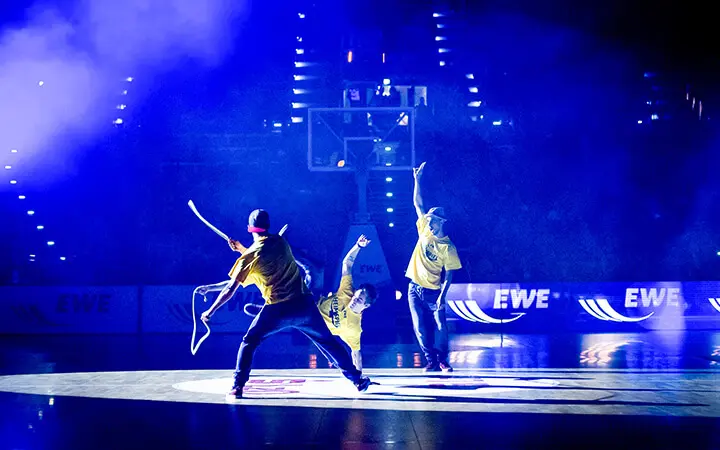 Drei Tänzer in gelben Hemden treten unter dramatischer blauer und violetter Beleuchtung auf einem Basketballplatz auf und schaffen eine elektrisierende Unterhaltungsszene mit Scheinwerfern und einem Basketballkorb im Hintergrund.