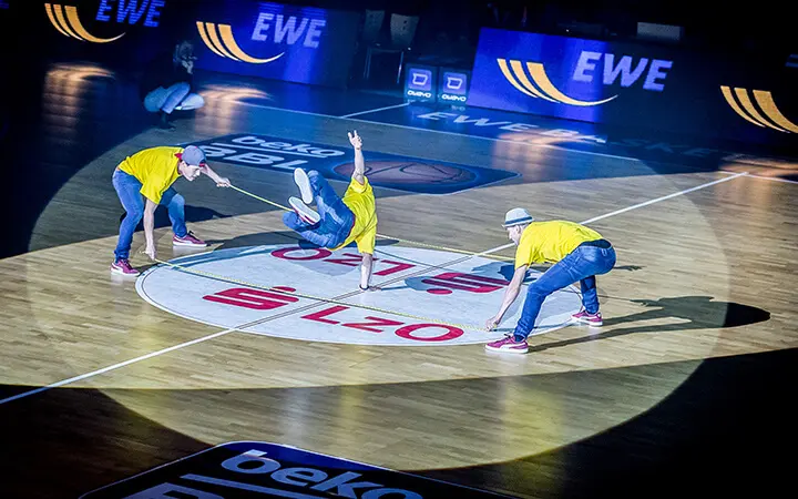 Drei Tänzer in gelben Hemden und Hüten zeigen auf einem Basketballfeld unter hellen Scheinwerfern energiegeladene Breakdance-Moves und sorgen so für ein elektrisierendes Unterhaltungsspektakel inmitten von EWE- und anderen Logos, die auf dem Boden und den Bildschirmen zu sehen sind.