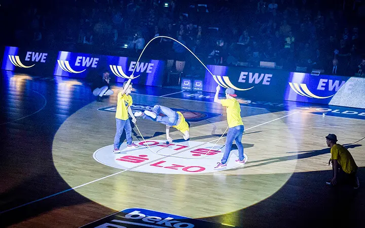 Drei Personen führen auf einem Basketballplatz unter einem Scheinwerferlicht eine energiegeladene Seilsprungnummer auf und sorgen für hochfliegende Unterhaltung, wenn einer in der Luft einen Sprung macht, zwei das Seil drehen und eine vierte Person in der Nähe hockt.