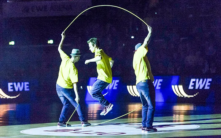 Drei Personen in gelben Hemden und Jeans führen auf einer beleuchteten Bühne eine unterhaltsame Seilsprungübung vor; zwei halten das Seil, während einer in der Mitte springt. Der Hintergrund ist dunkel, nur die beleuchteten "EWE"-Schilder sind zu sehen.