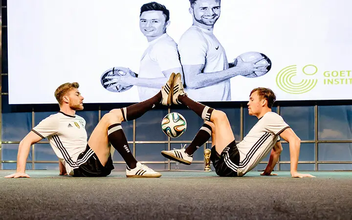 Zwei Männer in passenden Fußballtrikots sitzen sich auf dem Boden gegenüber und balancieren einen Fußball zwischen ihren Füßen in einer Art Fußball-Kür. Ein großer Bildschirm hinter ihnen zeigt die beiden, wie sie Fußbälle halten und lächeln.