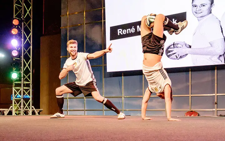 Zwei Personen in Sportkleidung zeigen auf der Bühne eine Fußball-Kür; die eine nimmt eine dynamische Pose ein, während die andere einen Handstand mit einem Fußball macht. Auf einer großen Leinwand hinter ihnen ist ein Schwarz-Weiß-Foto und der Name René zu sehen.