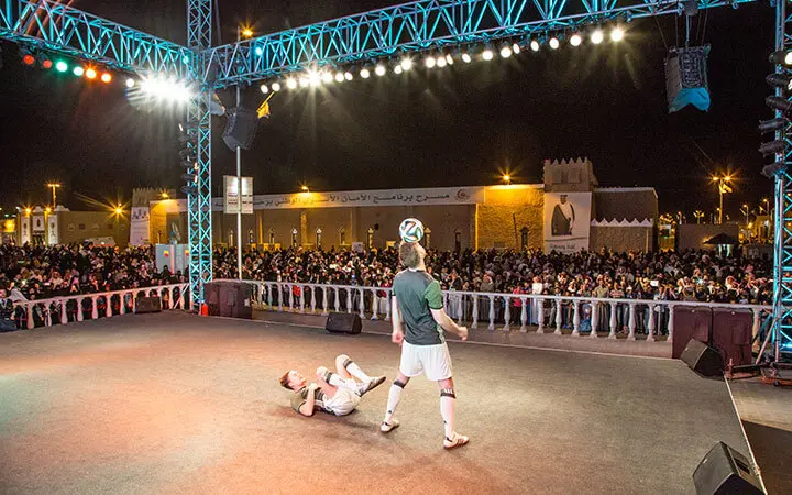 Zwei Künstler zeigen nachts auf einer hell erleuchteten Bühne beeindruckende Fußball-Freestyle-Tricks - einer balanciert einen Ball auf dem Kopf, während der andere auf dem Boden liegt. Eine große Menschenmenge schaut hinter einer Absperrung zu.