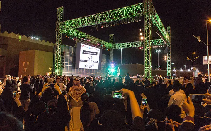 Eine große Menschenmenge versammelt sich nachts vor einer hell erleuchteten Freilichtbühne mit einem großen Bildschirm, auf dem Texte angezeigt werden. Viele Menschen machen Fotos oder Videos mit ihren Handys, während die Football-Freestyle-Künstler die lebhafte, festliche Veranstaltung anheizen.