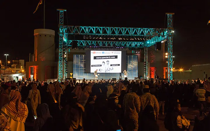 Eine große Menschenmenge versammelt sich nachts vor einer beleuchteten Freiluftbühne, auf der Künstler Fußball-Freestyle vorführen. Auf einem großen Bildschirm über der Bühne werden eine Website und ein Bild angezeigt, während einige Zuschauer traditionelle nahöstliche Kleidung tragen.