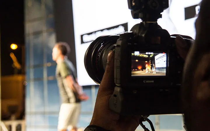 Nahaufnahme einer Kamera, die einen Fußball-Freestyler auf der Bühne mit einem großen, hellen Bildschirm im Hintergrund einfängt; die Person erscheint unscharf im Hintergrund, während das Kameradisplay ihre Tricks deutlich zeigt.