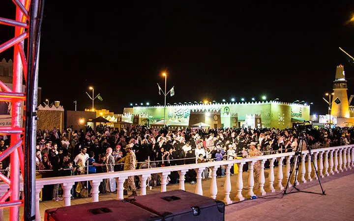 Eine große Menschenmenge versammelt sich nachts vor einem traditionell gestalteten Gebäude mit arabischer Beschriftung, das warm beleuchtet und dekoriert ist, während Fußball-Freestyler während eines kulturellen Festivals oder einer Veranstaltung im Freien auftreten.