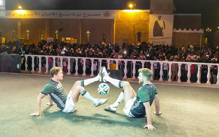Zwei Fußballspieler führen eine Fußball-Kür vor, bei der sie auf dem Boden sitzen und einen Ball mit ihren Füßen balancieren, während ein großes Publikum bei einer nächtlichen Veranstaltung im Freien zusieht und Fotos macht.