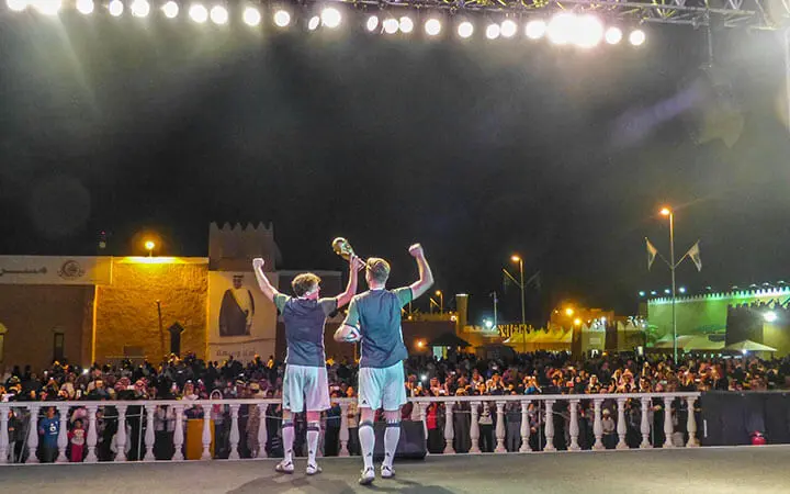 Zwei Fußball-Freistilspieler auf einer Bühne, die sich einer jubelnden nächtlichen Menge gegenübersehen, heben unter hellen Scheinwerfern einen Pokal und ihre Fäuste zum Jubel. Im Hintergrund sind Gebäude und Straßenlaternen zu sehen.