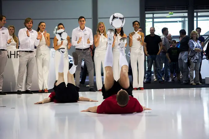 Zwei Fußballartisten liegen auf dem Rücken und jonglieren gekonnt Bälle mit den Füßen, während eine Gruppe weiß gekleideter Menschen in der Halle hinter ihnen steht und das Geschehen mit bewunderndem Klatschen verfolgt.