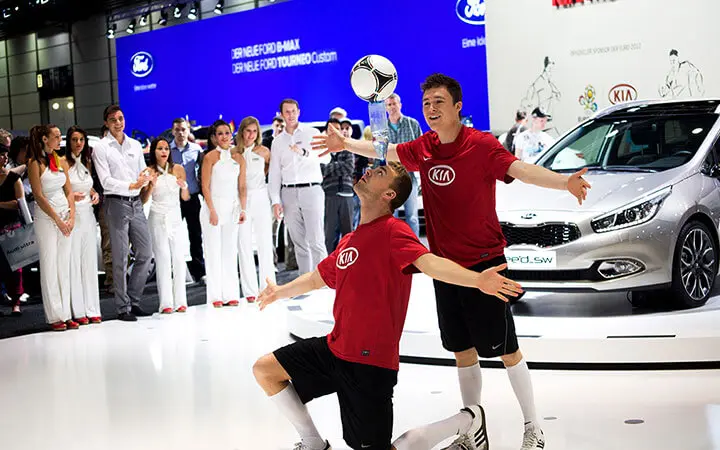 Zwei Fußballartisten in roten Kia-Trikots führen auf einer Autoshow einen Fußballtrick vor einem silbernen Kia-Auto vor, während eine Gruppe von Menschen in weißen Outfits im Hintergrund zuschaut und applaudiert.
