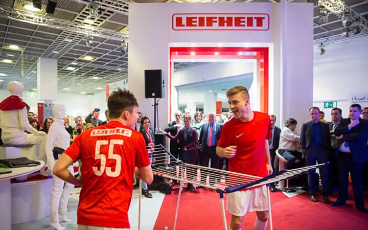 Zwei Männer in roten Sporttrikots spielen Tischtennis mit einem Wäscheständer als Netz, umgeben von einer lebhaften Menge bei einer Leifheit-Veranstaltung. Die spielerische Atmosphäre erinnert an eine Fußball-Show und wird von den Zuschauern begeistert bejubelt.