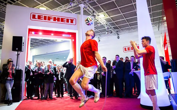Zwei Männer in roten Hemden zeigen eine dynamische Fußball-Show, indem sie in einem hell erleuchteten Messestand einen Fußball mit ihren Körpern jonglieren. Eine Menschenmenge beobachtet, wie der Stand stolz den Namen Leifheit in fetten roten Buchstaben über dem Eingang trägt.