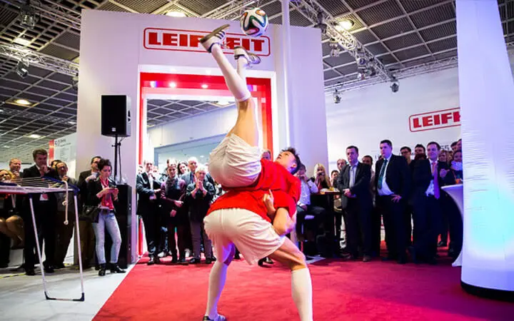 Zwei Fußballer in rot-weißen Trikots beeindrucken das Publikum bei einer Hallenveranstaltung mit einer akrobatischen Fußball-Show - einer balanciert kopfüber auf dem Rücken des anderen und hält einen Fußball gekonnt mit den Füßen fest.