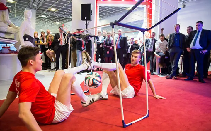 Zwei junge Männer in roten Hemden und weißen Shorts führen eine beeindruckende Fußball-Show auf, bei der sie auf einem roten Teppich im Sitzen ein Fußballspiel mit Tricks spielen, während eine Menge formell gekleideter Menschen zusieht.