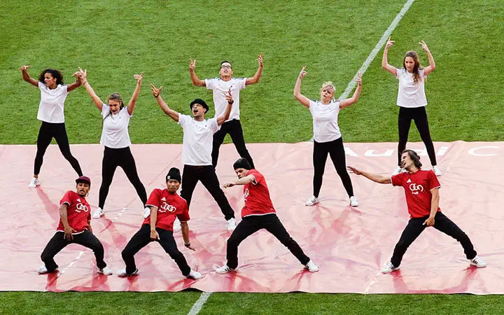 Eine Urban-Dance-Gruppe tritt auf einem Feld auf, mit fünf Tänzern in roten Hemden und schwarzen Hüten vor und fünf Tänzern in weißen Hemden hinter ihnen, die alle energiegeladene Posen auf einer roten Matte einnehmen.