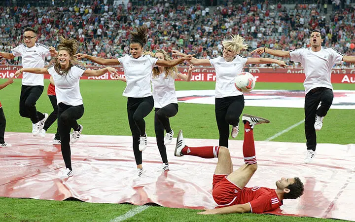 Ein Fußballspieler in rotem Trikot vollführt Tricks auf dem Rücken und balanciert einen Ball auf seinen Füßen. Hinter ihm springen Urban-Dance-Künstler in weißen Hemden mit ausgestreckten Armen, während eine große Menschenmenge im Stadion zusieht.