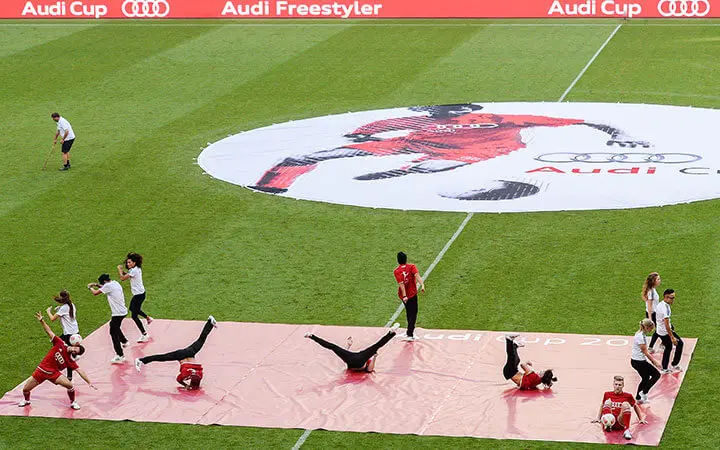 Eine Gruppe von Urban Dance-Performern belebt das Fußballfeld in der Nähe eines großen runden Banners mit einer Fußballergrafik während des Audi Cups. Einige Tänzer befinden sich mitten in der Bewegung auf einer roten Matte, während andere in der Nähe stehen.