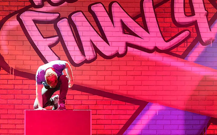 Eine Person kniet während einer Parkour-Show auf einer roten Plattform vor einer hell erleuchteten Backsteinmauer mit bunter Graffiti-Kunst und der großen Aufschrift FIN4Y. Rosa und lila Licht erzeugt einen dramatischen Effekt.