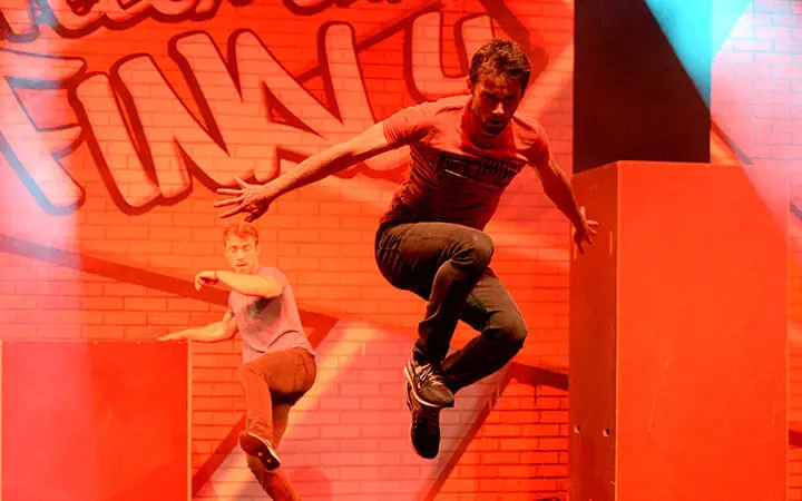 Zwei Männer befinden sich in der Luft und springen während einer dynamischen Parkour-Show auf einer hell erleuchteten Bühne mit Rottönen und Graffiti-Hintergrundgrafiken.