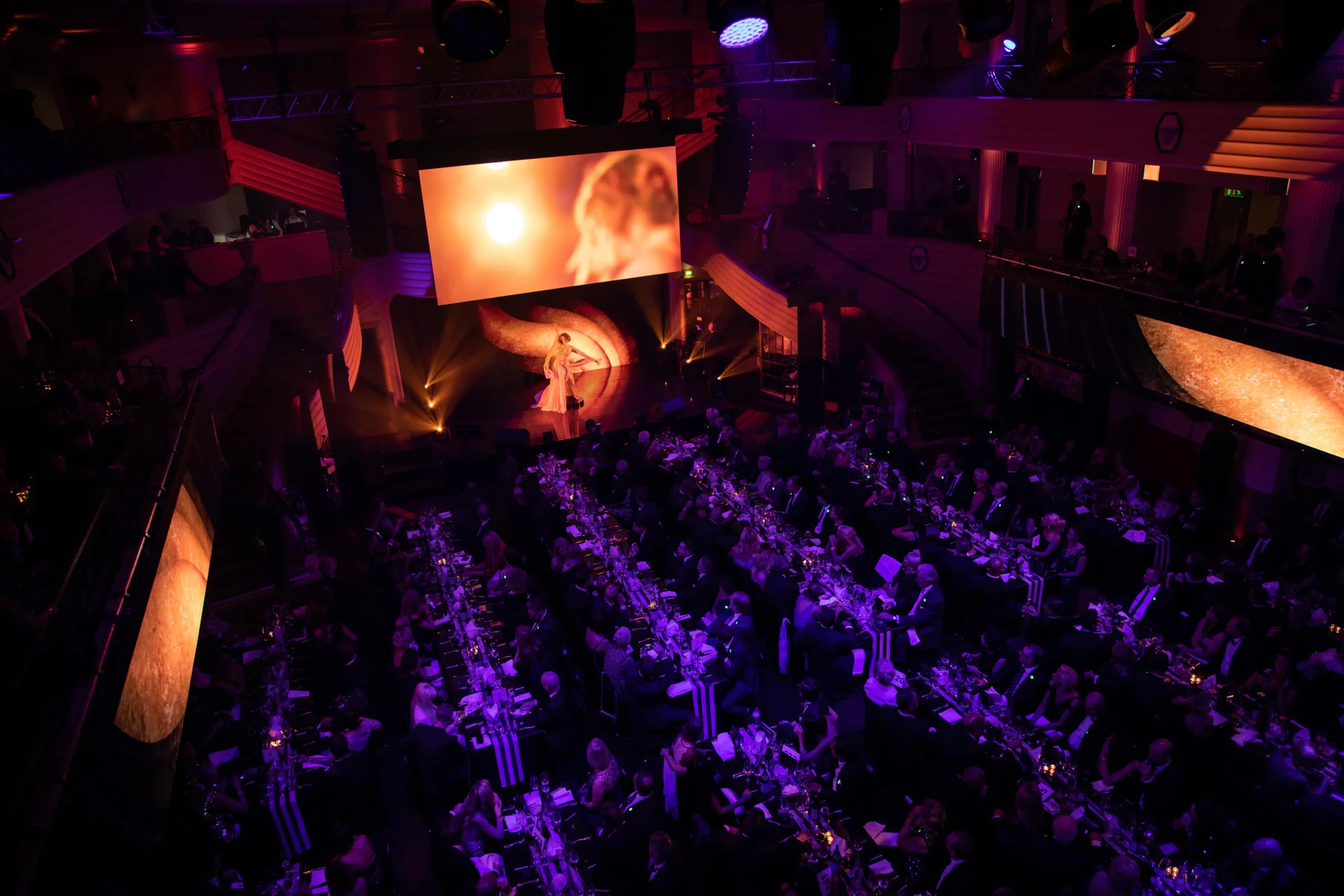 Ein großes Publikum sitzt an langen Tischen in einem schwach beleuchteten Saal mit violetter Beleuchtung und beobachtet die CIRCLE SHOW auf der Bühne, wo eine Person vor einem großen Bildschirm steht, auf dem ein orangefarbenes, abstraktes Bild zu sehen ist, das an das Branding von McDonald's erinnert.