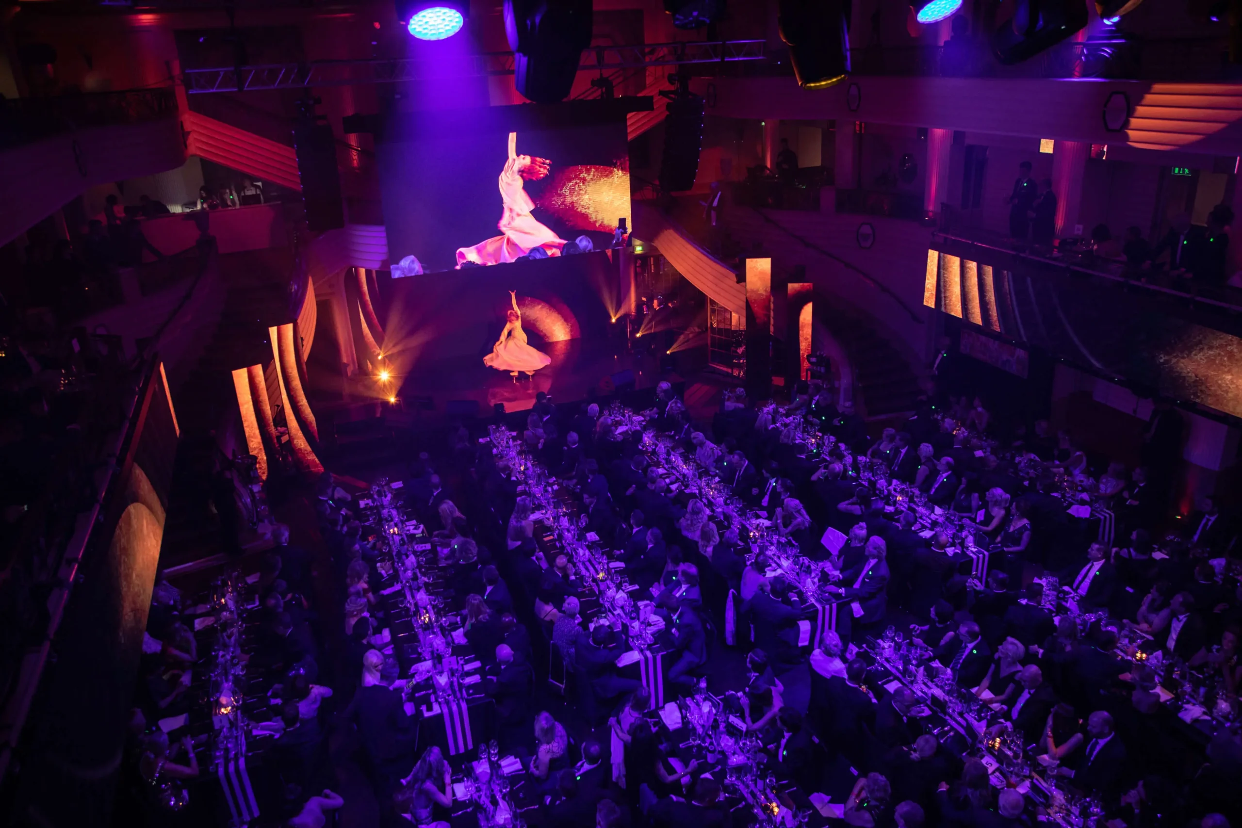 Ein großer, eleganter Bankettsaal ist mit Menschen gefüllt, die an langen Tischen unter lila und orangefarbener Beleuchtung sitzen. Auf der Bühne dreht sich ein Darsteller für die Mc Donalds - CIRCLE SHOW, über dem ein großer Bildschirm eine Nahaufnahme der Vorstellung zeigt.