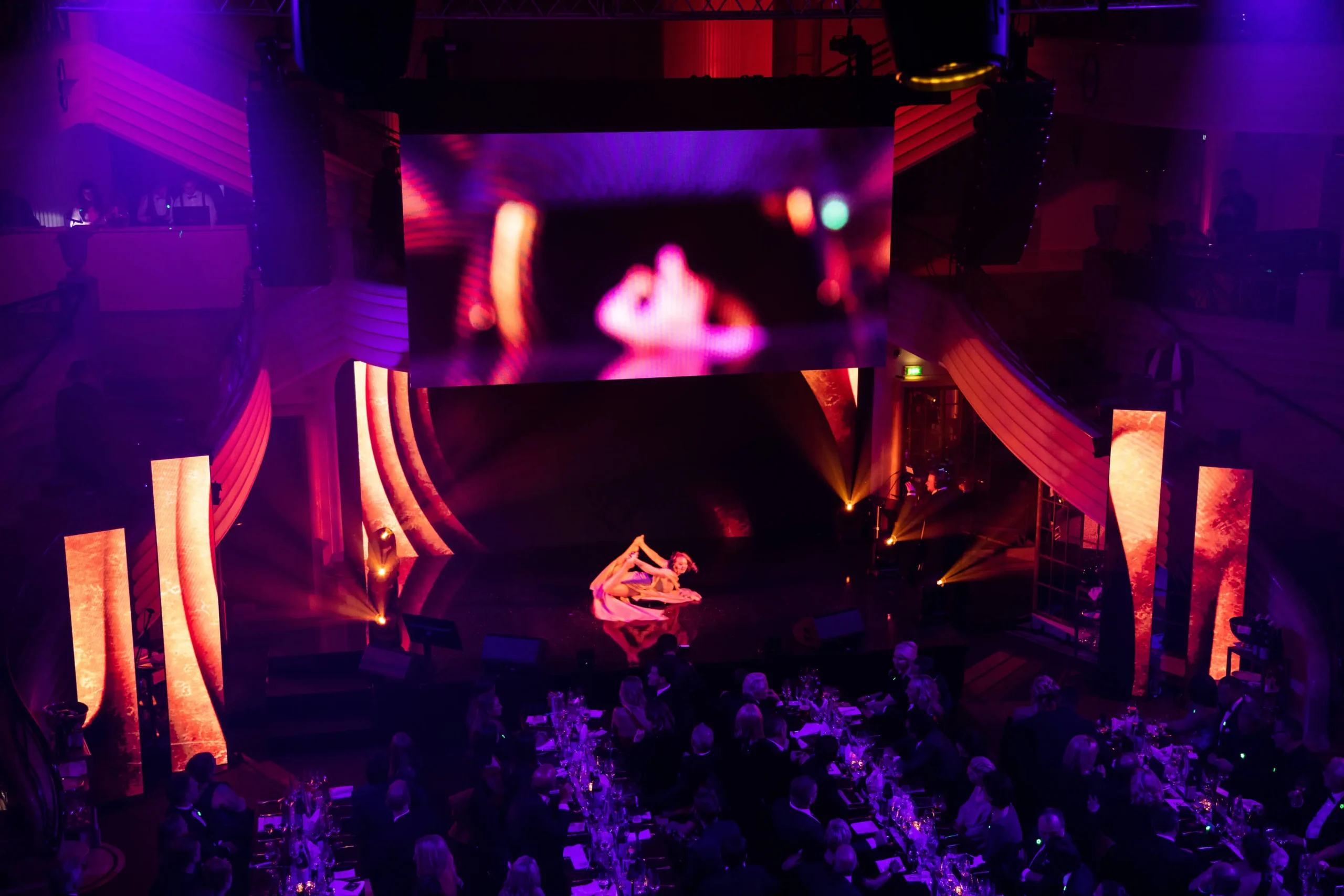 Auf einer schwach beleuchteten Bühne mit violetten und orangefarbenen Lichtern findet die Mc Donalds - CIRCLE SHOW statt, bei der zwei Tänzer ineinander verschlungen auf dem Boden unter einer großen, unscharfen Projektion tanzen, während die an Tischen sitzenden Zuschauer im Vordergrund aufmerksam zusehen.